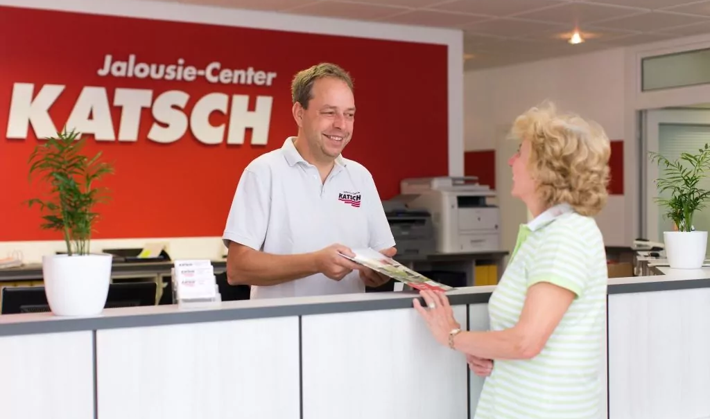 Ein Mitarbeiter des Jalousie-Center Katsch berät eine Kundin an einem Tresen vor einer Wand mit großem Logo.