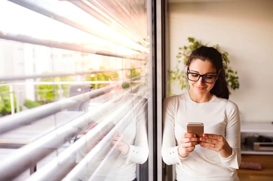 Modernes Einfamilienhaus mit Rollläden Frau steht an ihrem Fenster vor dem Rollladen und tippt an ihrem Handy